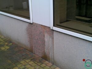 Gevel met granieten afwerking waarop roestvlekken en vochtsporen zichtbaar zijn onder een raamkader