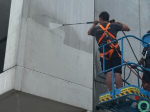 Een medewerker reinigt een vuile betongevel met een hogedrukreiniger vanuit een hoogtewerker.