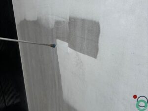 Een vervuilde betonnen wand wordt gereinigd met een hogedrukspuit, waardoor een duidelijk verschil zichtbaar is tussen schoon en vuil beton.