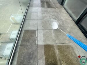 Vervuilde terrastegels worden met een hogedrukreiniger gereinigd, met duidelijk zichtbaar kleurverschil tussen het vuile en het schone oppervlak.