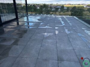 Pas gereinigd balkon met natte tegels die glanzen in het zonlicht, met zicht op een open landschap en moderne glasbalustrade.